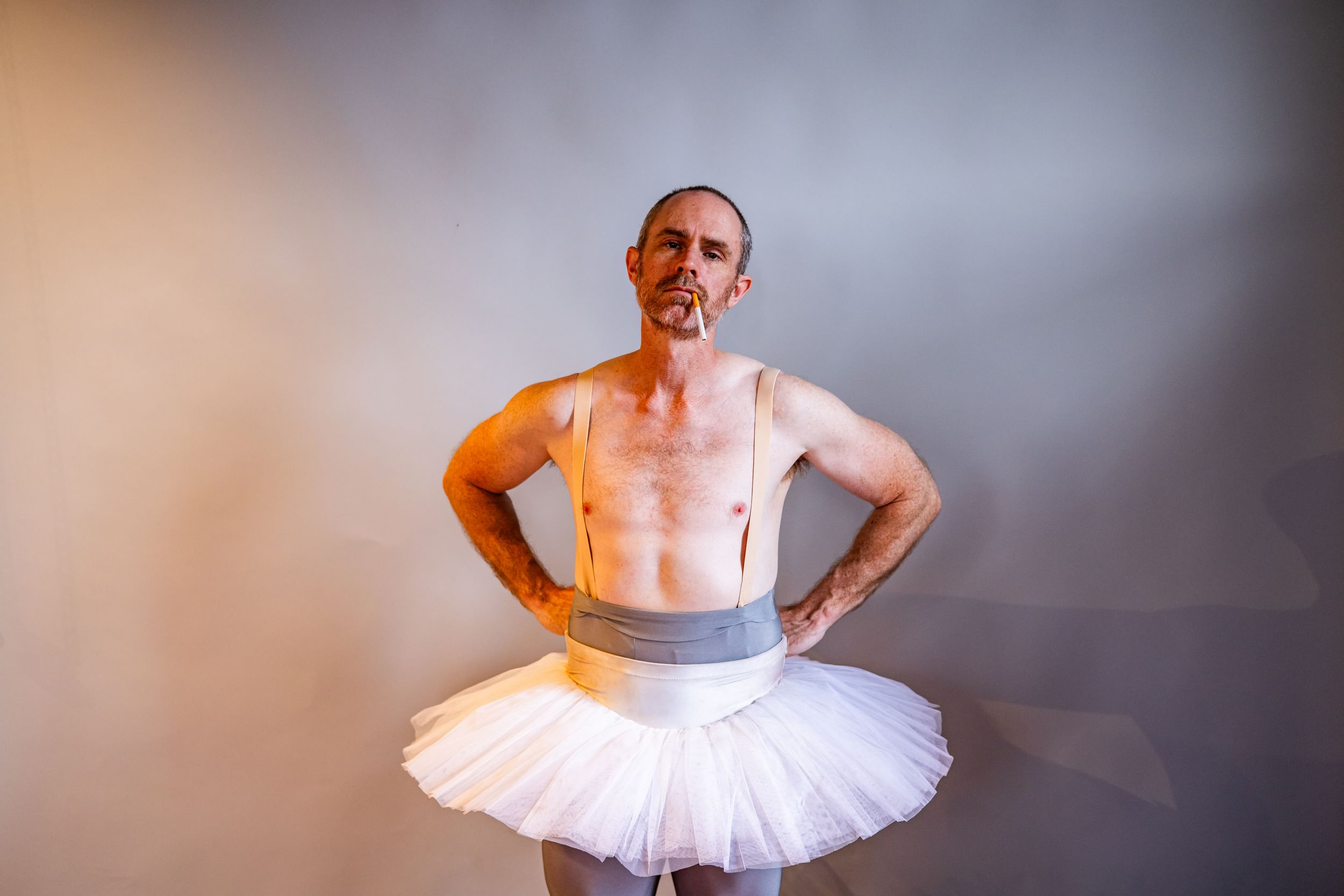 Davis, shirtless, with nude suspenders & white tutu, stands with hands on hips, cigarette in his mouth.