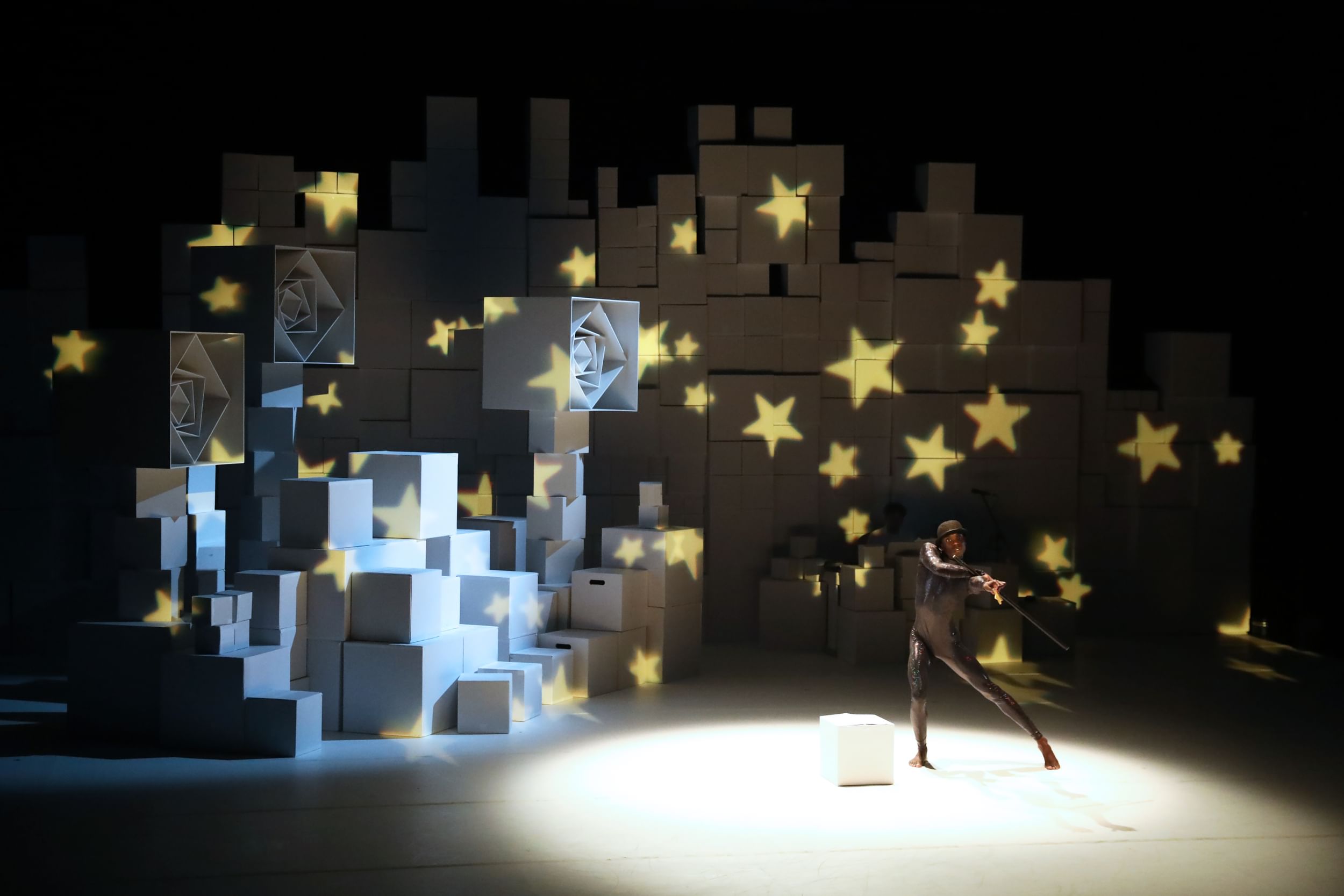 A performer in an all black fencer fit, colorful box shaped items stacked on stage, with stars decorating the backdrop