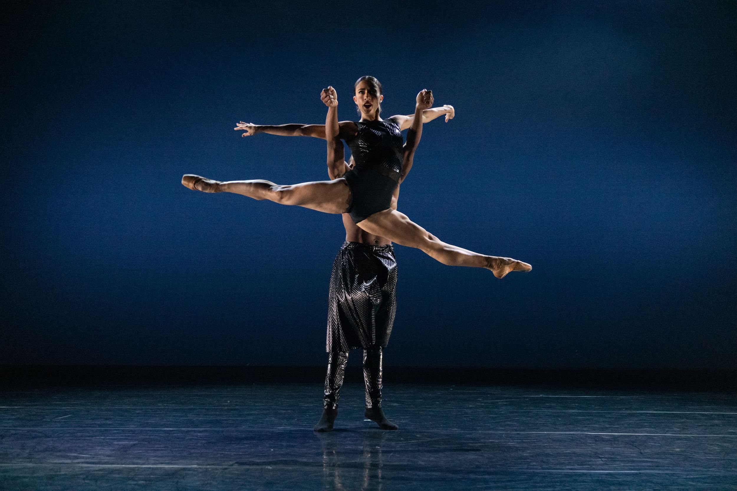 Two dancers on stage in pas de deux, one lifting the other dancer as they do a split in the air