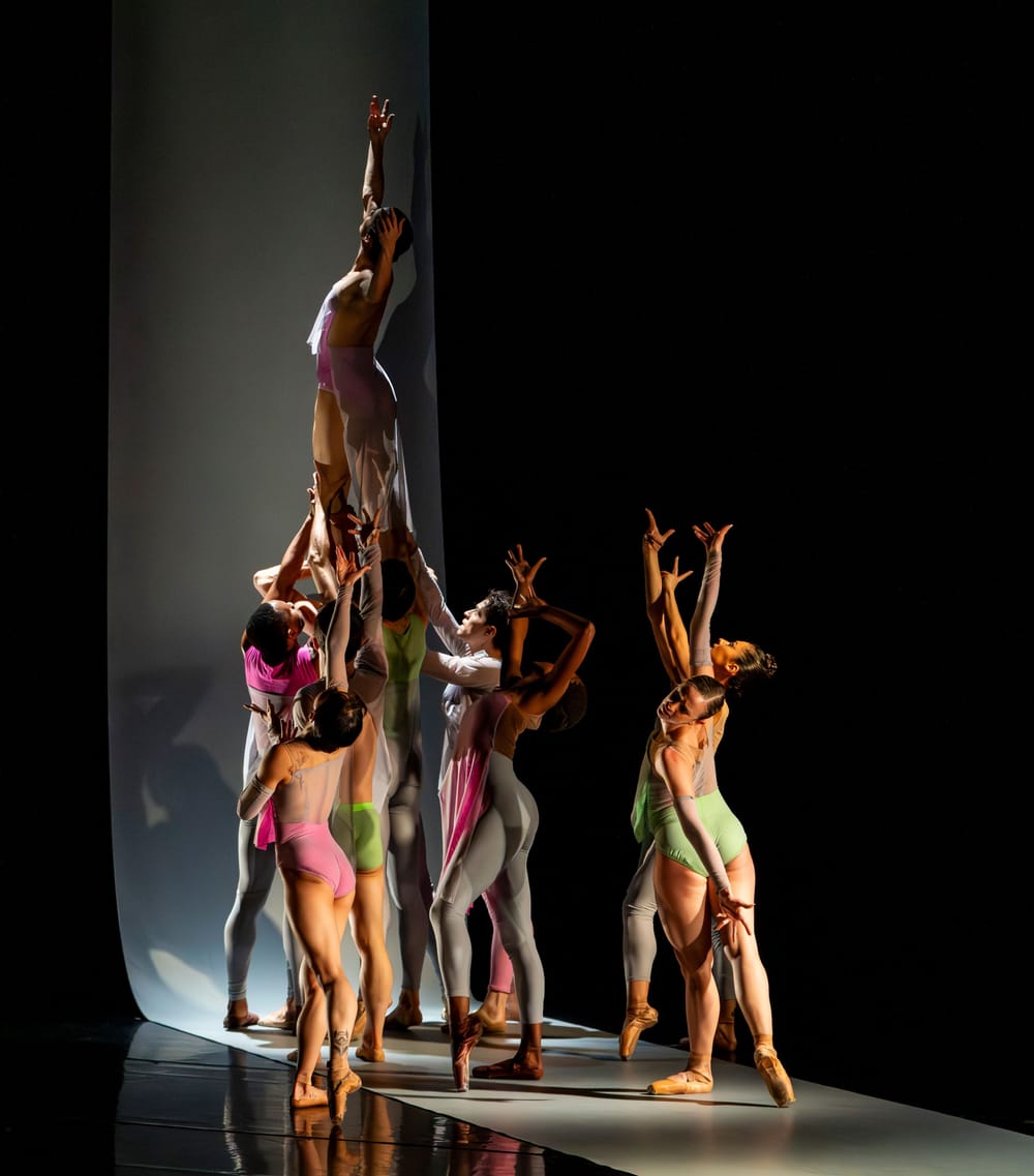 Eight dancers in gray, pink & green pose reaching upward, some of them holding a female dancer vertically aloft.