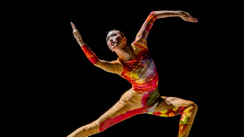Kim dances with her arms & legs all at different angles, wearing a striking bodysuit with warm, bright colors.