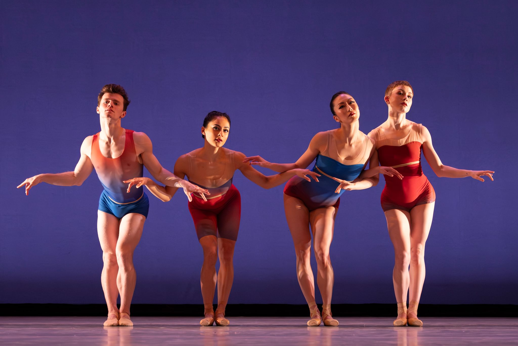 The dancers, in sheer leotards, stand in a line & lean forward with hips thrust out, arms bent to create a graceful wavy line