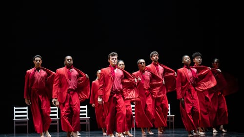 About 12 dancers in loose, bright-red costumes strike the same pose, pulling one side of their red blazers out with faces up
