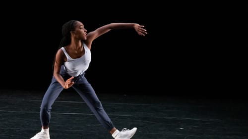 Black dancer in athletic wear & white sneakers in a contorted pose: her arms & legs out, palms & feet in different directions