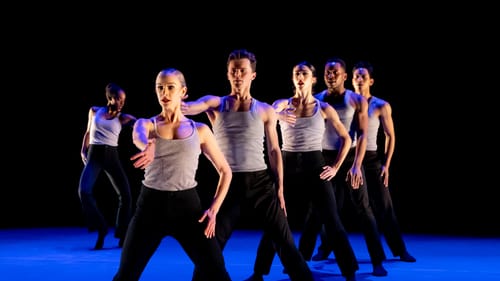 Line of 5 dancers in a similar pose, facing front with legs spread. 1 dancer alone at left They wear black pants & gray tanks