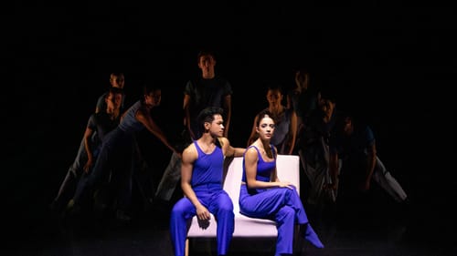 In sleeveless blue costumes, 2 dancers sit on a white couch, Palazo with his arm on the back, Forcella turning away from him