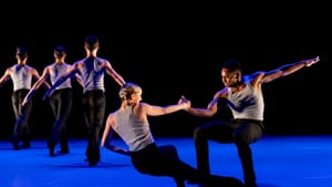 3 dancers seen from behind at left on a blue-lit stage; a male/female duet balance each other, connected by one clasped fist