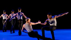 3 dancers seen from behind at left on a blue-lit stage; a male/female duet balance each other, connected by one clasped fist