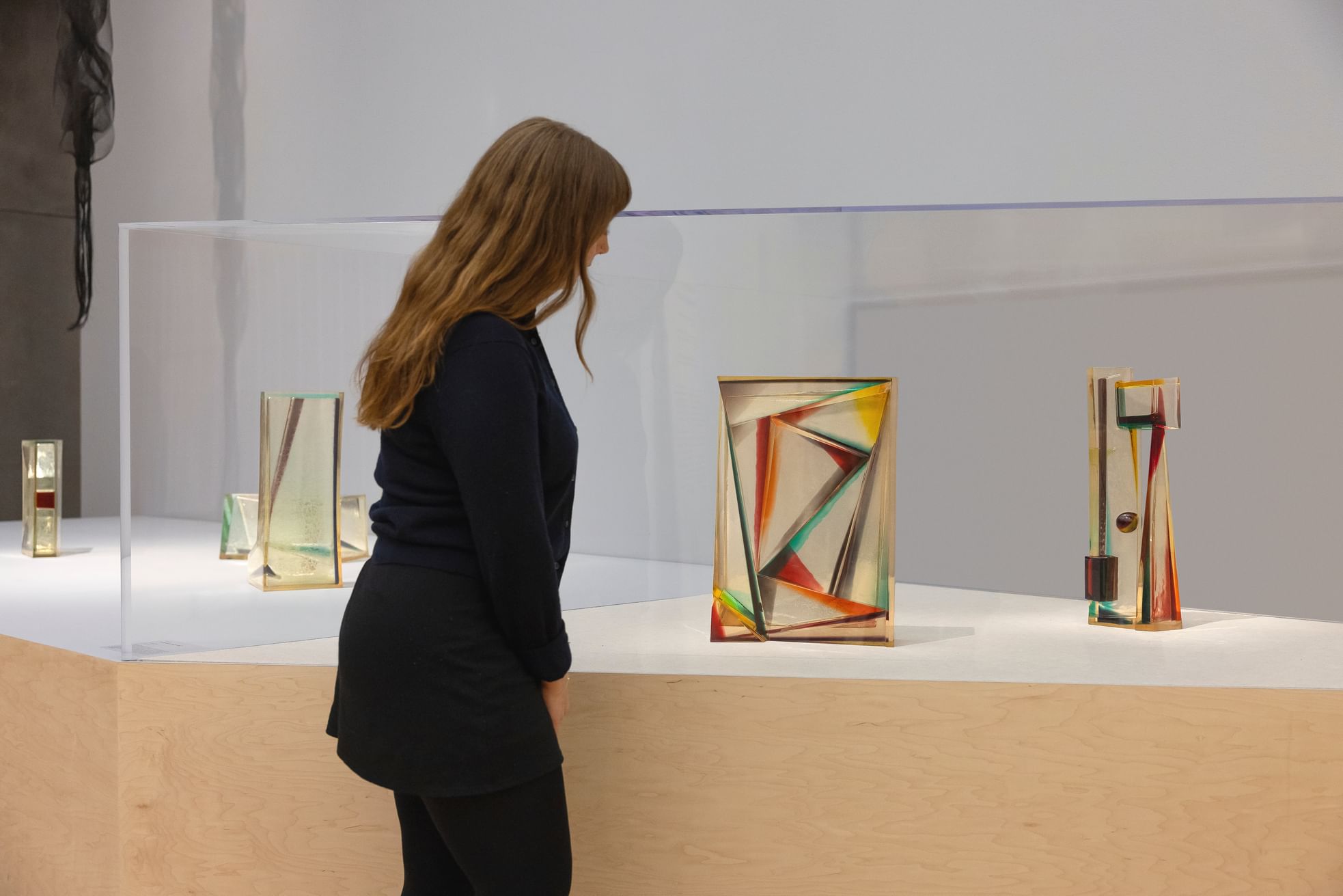 A woman with long brown hair looks closely at a series of colored glass sculptures with delicate lines and shapes.