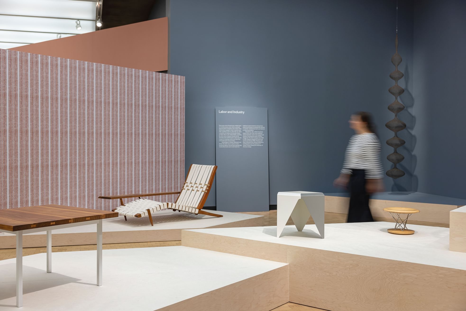 A motion-blurred person walks through a gallery of small, elegant furniture items with minimalist midcentury modern vibes.