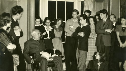 Black & white vintage photo of Barnes speaking to about 15 students, sitting casually in a chair with a small dog on his knee