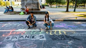 Two people squat by chalk writing on asphalt that says 'Juneteenth' 'Barnes on the Block.'