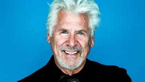 Portrait photo of Bostwick. Arms crossed, smiling at camera. Wears all black, with gray hair. Blue backdrop