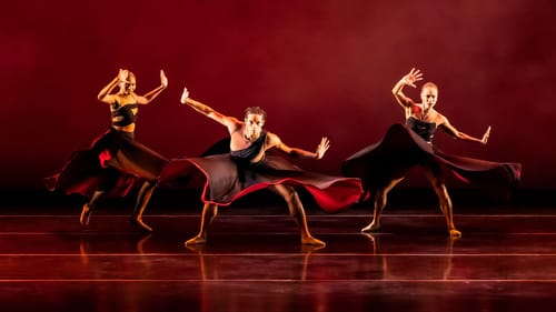 Three dancers pose, their outfits flowing out as they are captured mid-move. A foggy black and red background behind them.