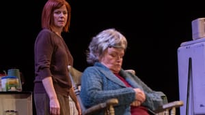 Quinn, a red-haired woman in brown, looks resentfully at Walsh, an older woman dozing in a rocking chair in a dingy kitchen