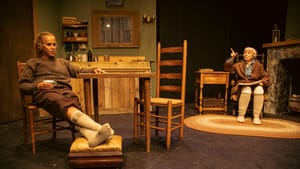 Maureen & Mag sit in a rustic living room with wood furniture. Mag looks accusing, Maureen tired, her feet on a footrest.