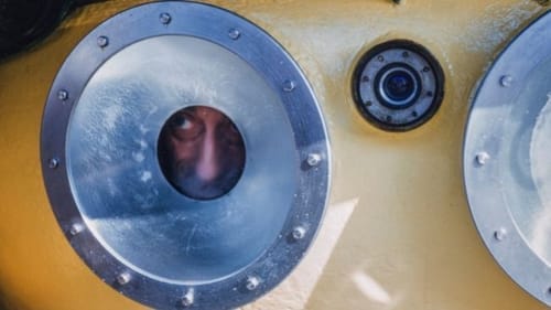 A close-up on the outside of a yellow submarine. A man’s face peeks curiously out of a small, metal-rimmed porthole window.