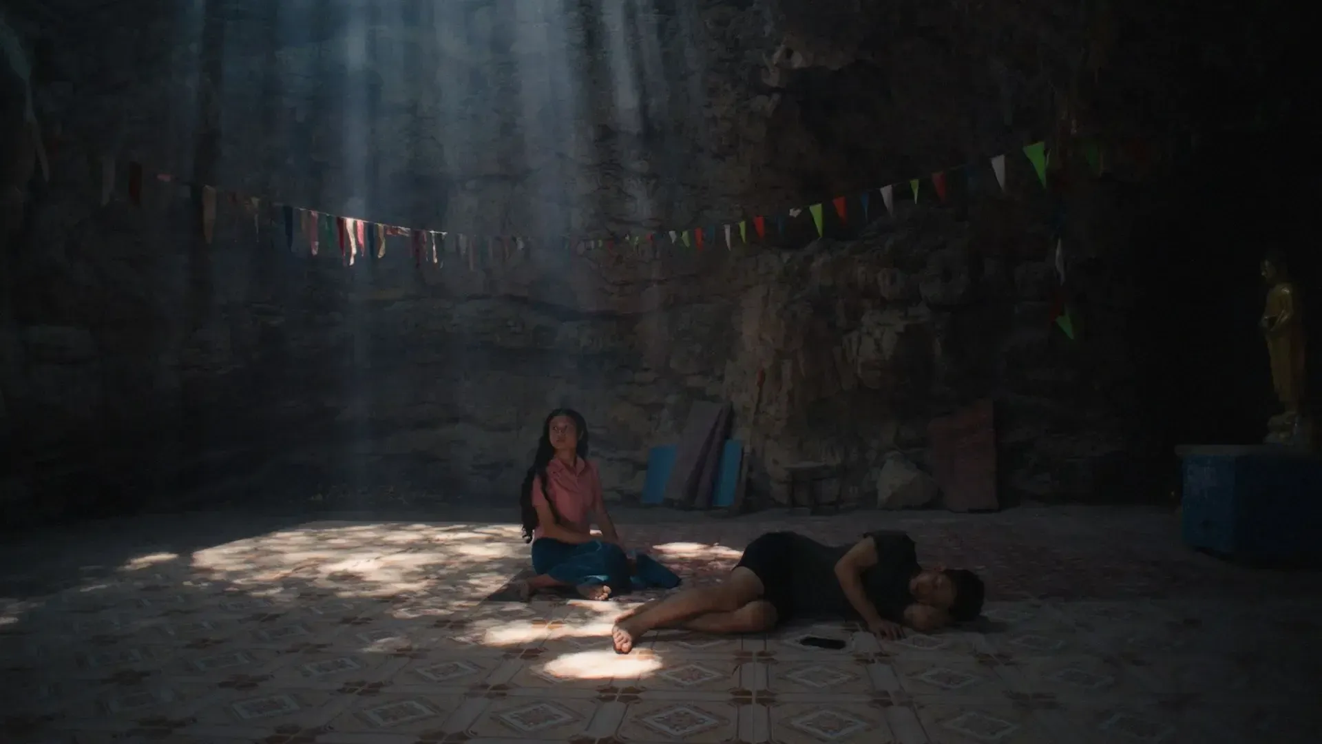 A girl sits next to a woman who lies on the ground, in what looks like a cave with a textured, patterned floor and a statue