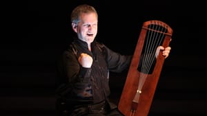 Bagby, a white man with short gray hair, performs in black with his hand-held wooden 6-string harp.