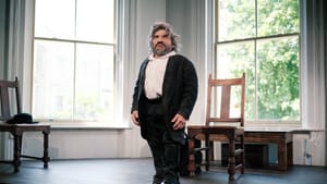 Povinelli, a little person with wavy gray hair and beard, stands proudly on a windowed stage, wearing a black suit.