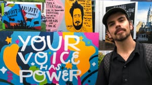 Benner, a man in his late 30s in a black button-down, smiles proudly while pointing to a mural that says your voice has power