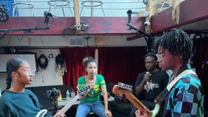 Four children/young adults in a studio. 2 play guitar, two hold mics. Camera equipment pointing at the guitar players