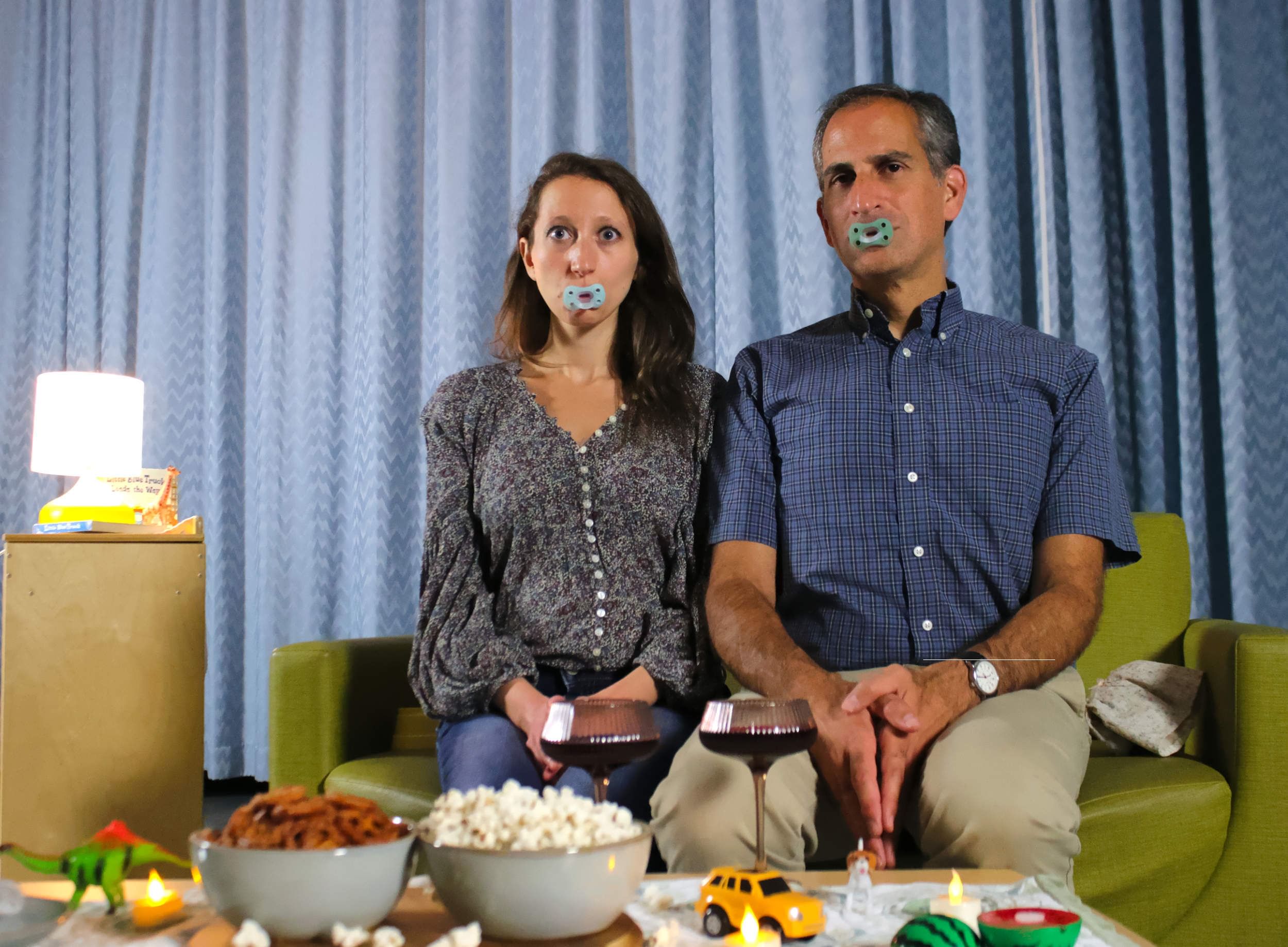 Cylinder & Barg sit straight-backed on a green couch, pacifiers in their mouths, snacks and toys on the coffee table.