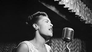 B&w photo of Billie Holiday. On stage, she sings in front of a vintage microphone