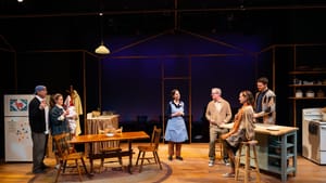 6 actors in contemporary clothes on a set like a suburban kitchen make a toast to Lamm at center, wearing a blue jumper