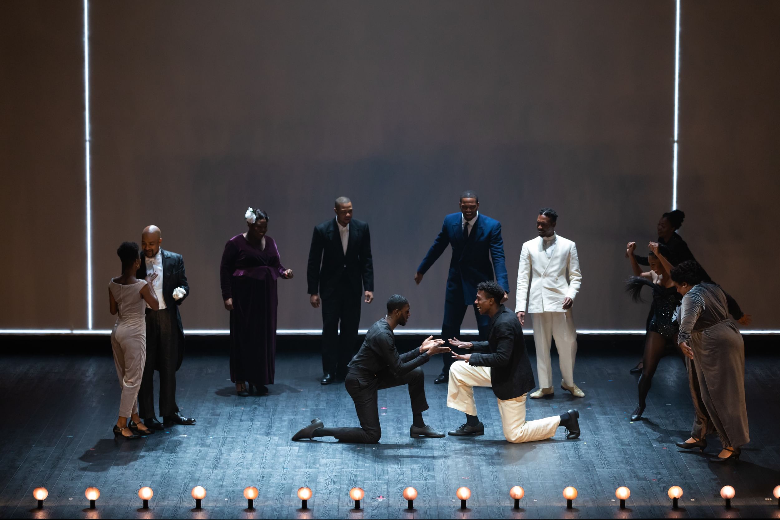About a dozen Black performers in formalwear of various styles, colors smile towards two people centered, facing each other