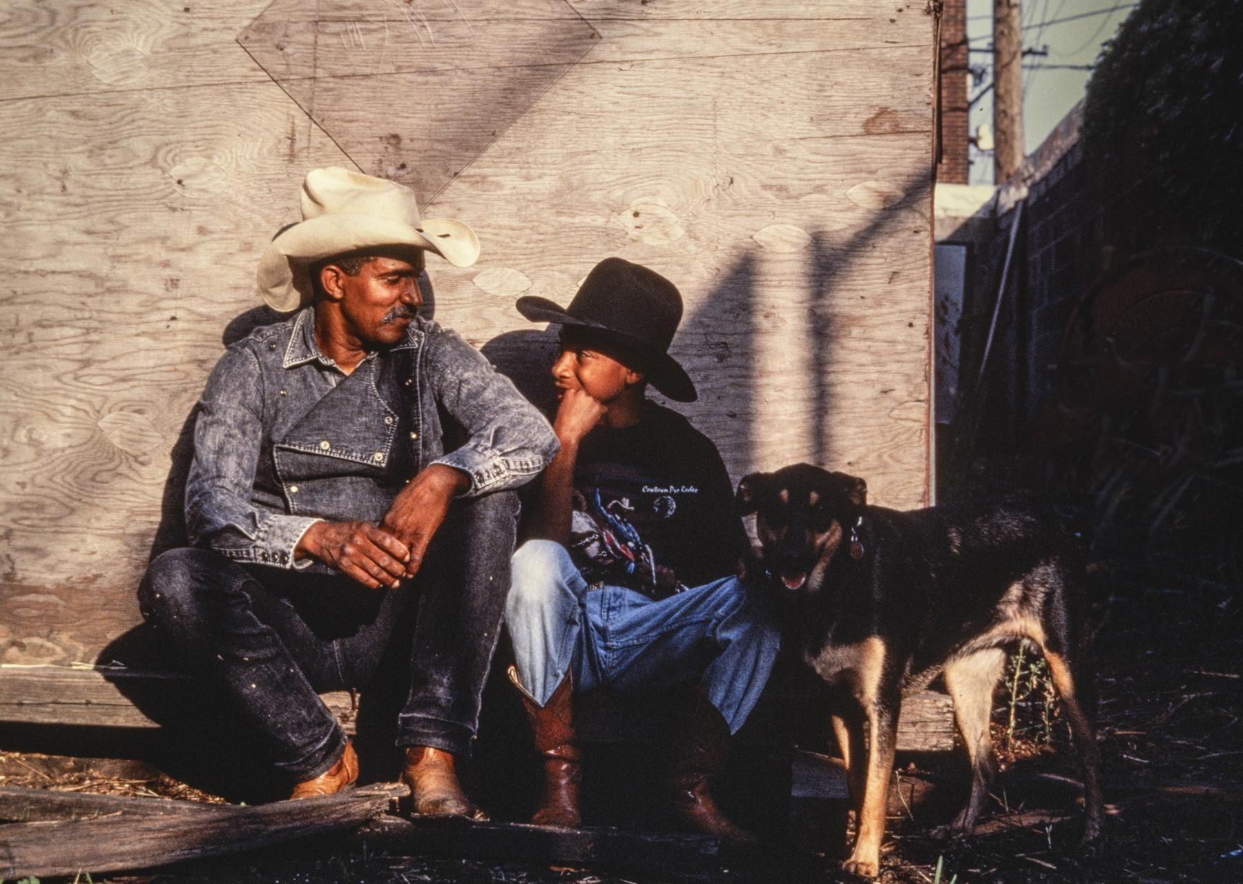 A Black man with a gray mustache sits companionably with a Black boy. They wear denim, cowboy hats, and cowboy boots.