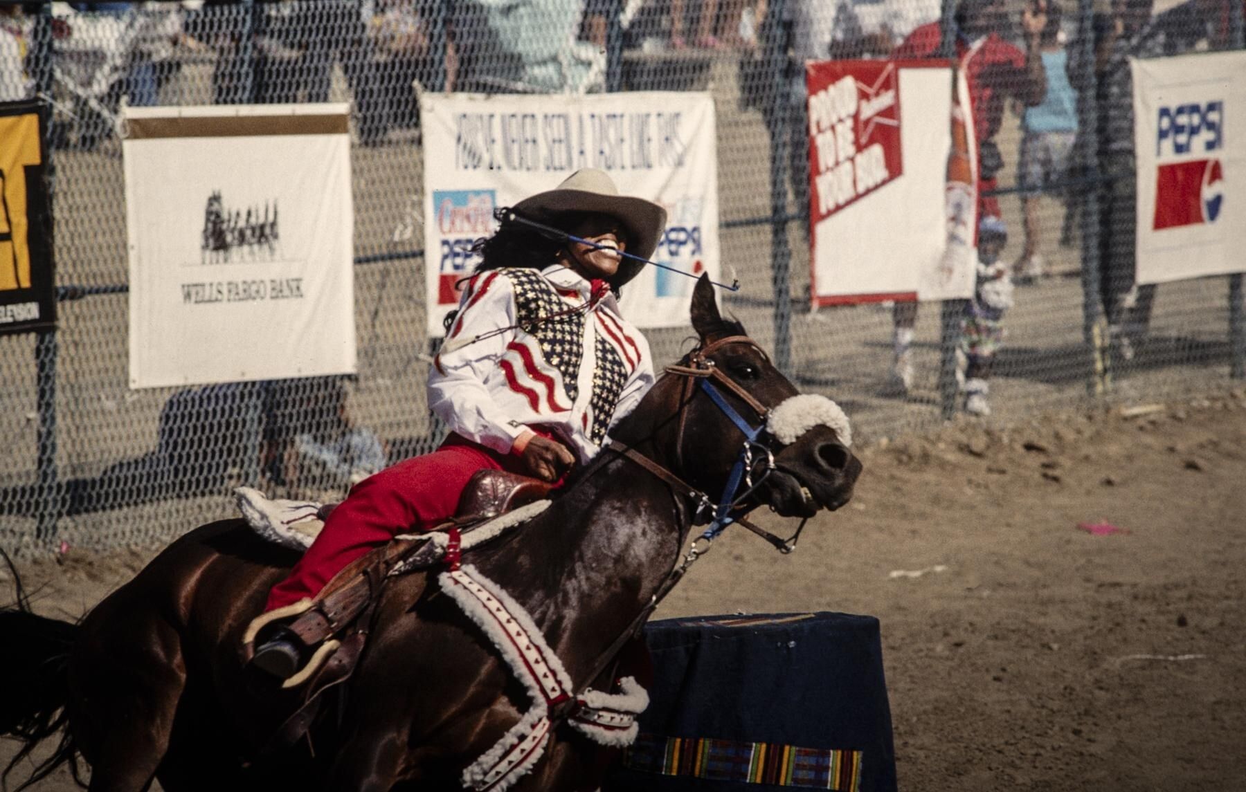 A Black woman in a cowboy hat and American-flag outfit, a riding crop in her teeth, dashes around a tight corner on horseback