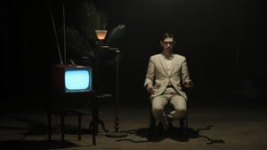 A person dressed in a beige suit sits in a chair by a CRT TV with a white screen, a plant in the dimly lit background