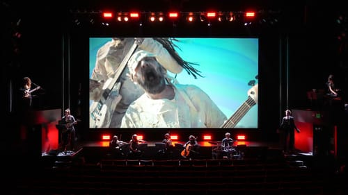 Rock musicians in white howl on the big screen; Timur and 8 musicians, lit in red, line the stage in front of the film.