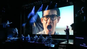 Close-up on Timur’s open-mouthed singing face on the big screen; Timur singing live with 8 musicians in front, lit in blue