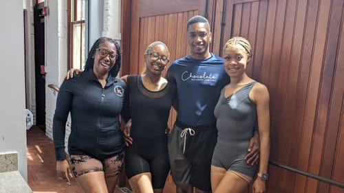 The artists pose smiling in rehearsal clothes with their arms around each other, in a sunny studio with large wooden doors.