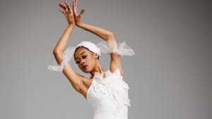 Sitting on a rectangular block at an angle with ankles crossed, Barnwell poses in a white tutu with arms gracefully raised