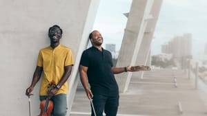 Two Black men pose outside, laughing, holding their violins