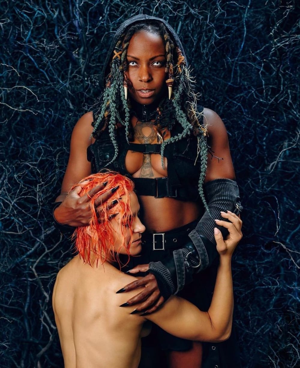 A light-skinned person submissively, sensually embraces a dark-skinned person in a strappy black ensemble & foreboding face