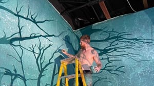 Montana on a yellow ladder, with no shirt and paint-splattered shorts, paints tree branches on the wall of the set.