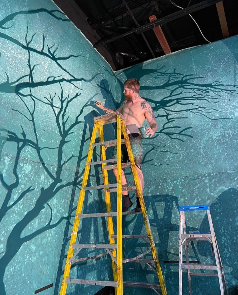 Montana on a yellow ladder, with no shirt and paint-splattered shorts, paints tree branches on the wall of the set.