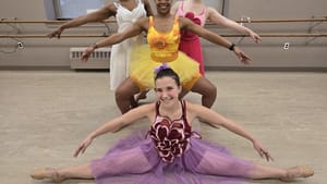 Four dancers in tutus in different colors, designs, pose for a group photo, one dancer in a split in front of other three