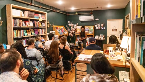 Morgan, a Black writer in black clothes and glasses, addresses a small crowd of people seated in a small bookstore.