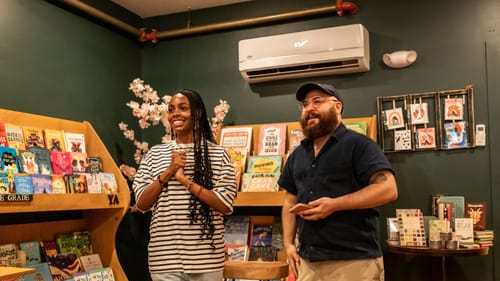 Townes, a Black woman in a striped tee, poses smiling with Shendelman, a bearded white man, in front of the bookstore crowd