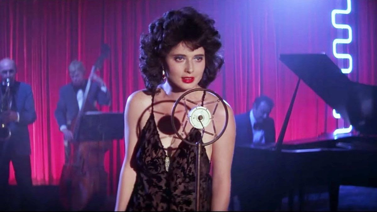 Rossellini, with a curly black 80s coif and evening dress, sings into a vintage mic at a nightclub.