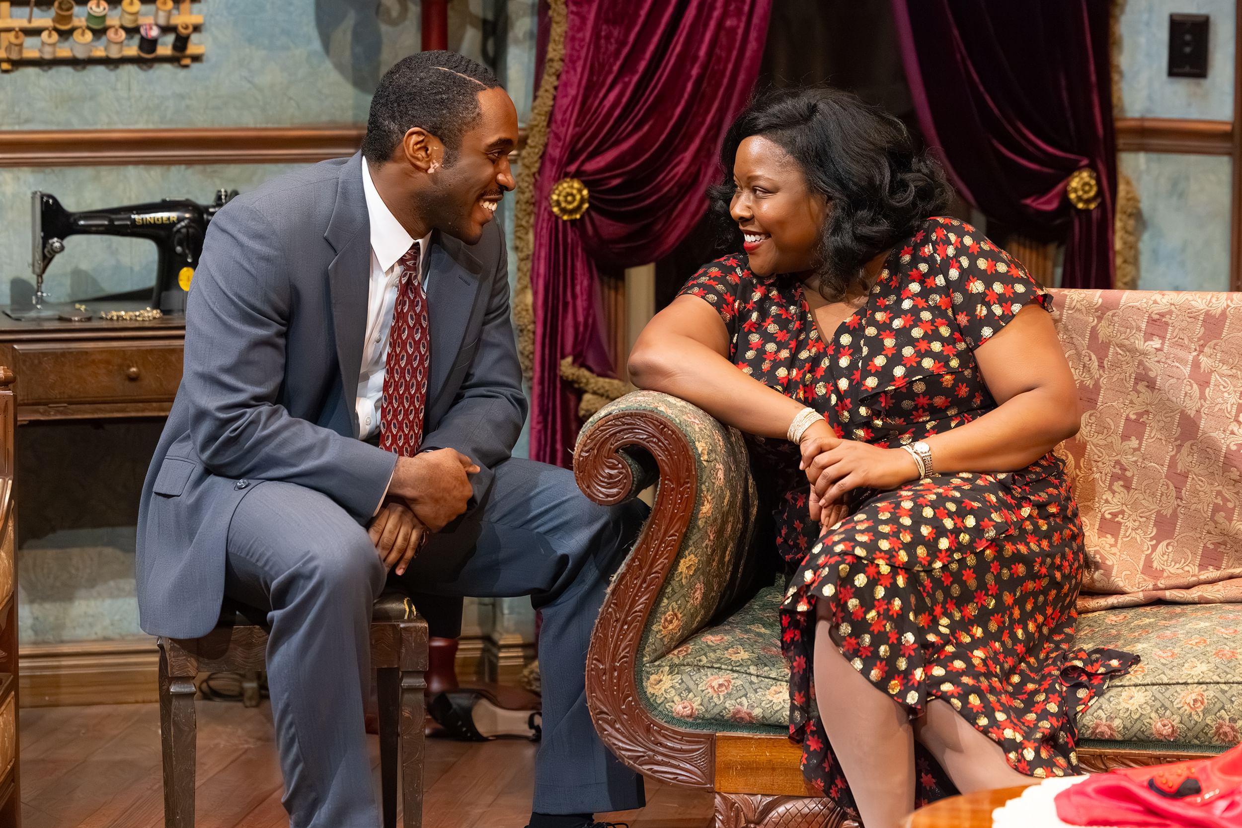 Kanu & Pullum smile flirtatiously at each other in a 1930 living room, a maroon velvet curtain draping the door behind them