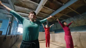 Three dancers in matching solid color shirts and sweats stretch out their arms, standing in an empty pool area