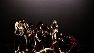 Dancers, all masked, in casual clothes, on dim stage. One dancer is on the floor, others stand over with their hands out over