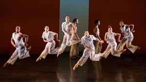 6 dancers in roomy sheer white outfits form a zigzag in the air with their bodies, all leaping in the same angular pose.