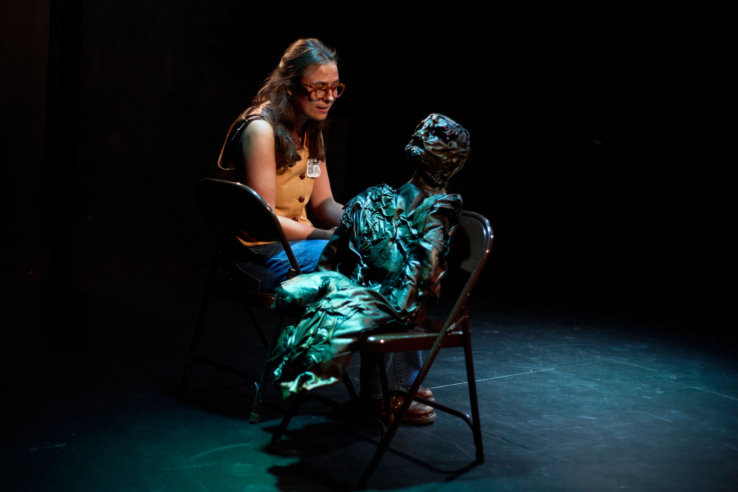 Waterman, a white woman with glasses & brown hair, sits next to a creepy black puppet that looks like a tortured mummy